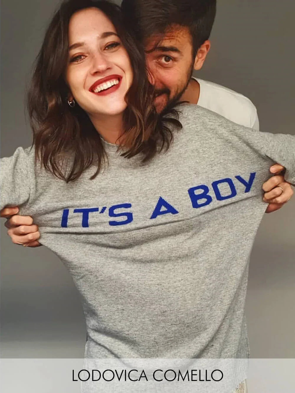 Donna con maglia premaman grigia con scritta blu 'IT'S A BOY' e uomo alle spalle.