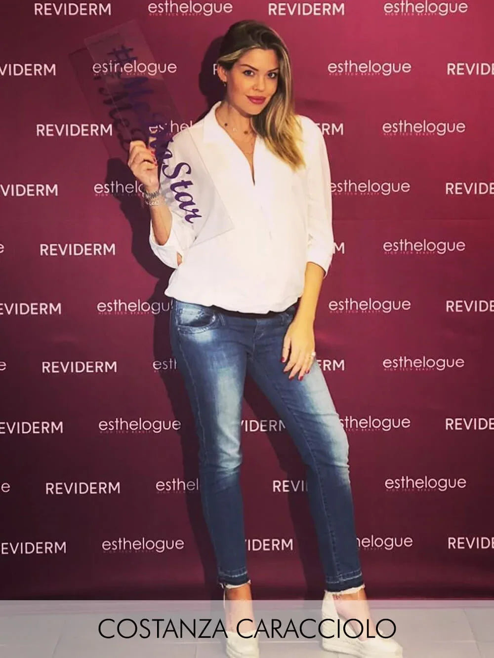 Donna con camicia bianca e jeans premaman blu davanti a sfondo viola.