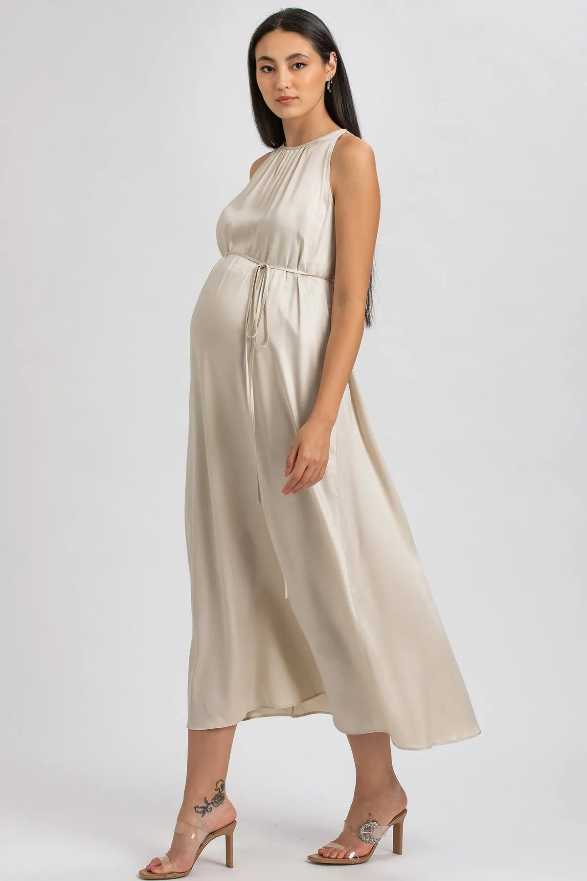 Donna incinta con abito premaman elegante beige in raso, sandali con tacco, fondo neutro