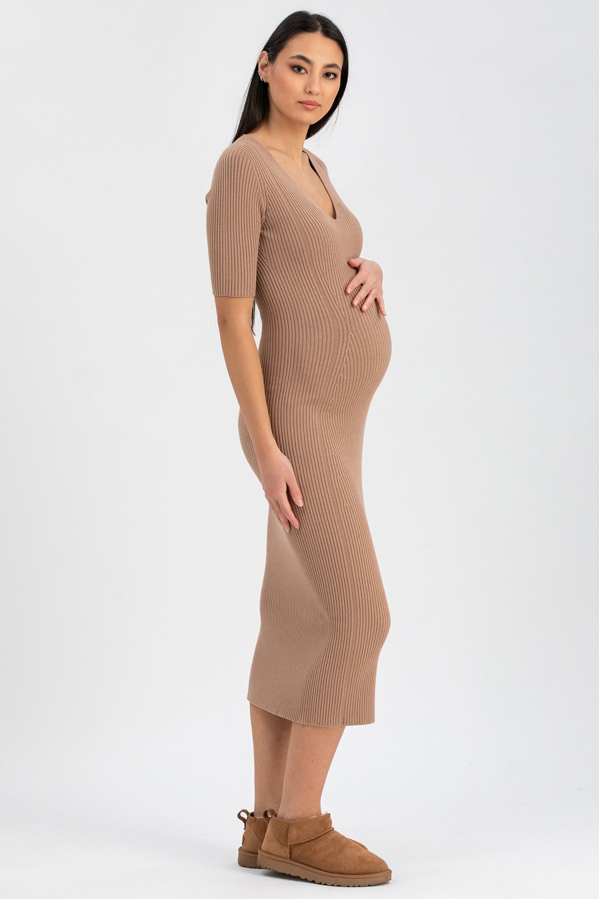 Donna incinta con abito premaman in maglia beige a coste e stivaletti, posa su sfondo neutro.