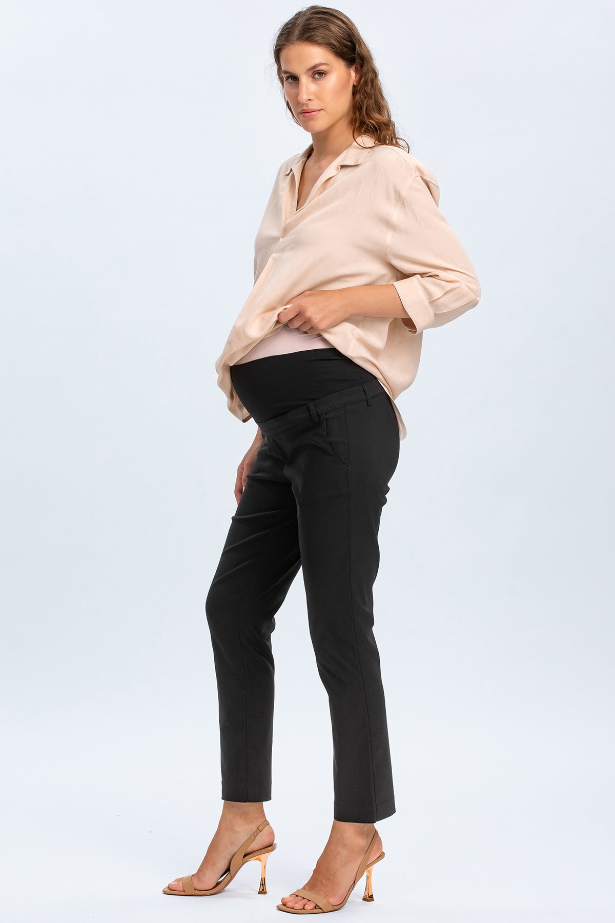 Donna incinta con camicia beige e pantaloni premaman neri eleganti, su sfondo chiaro.