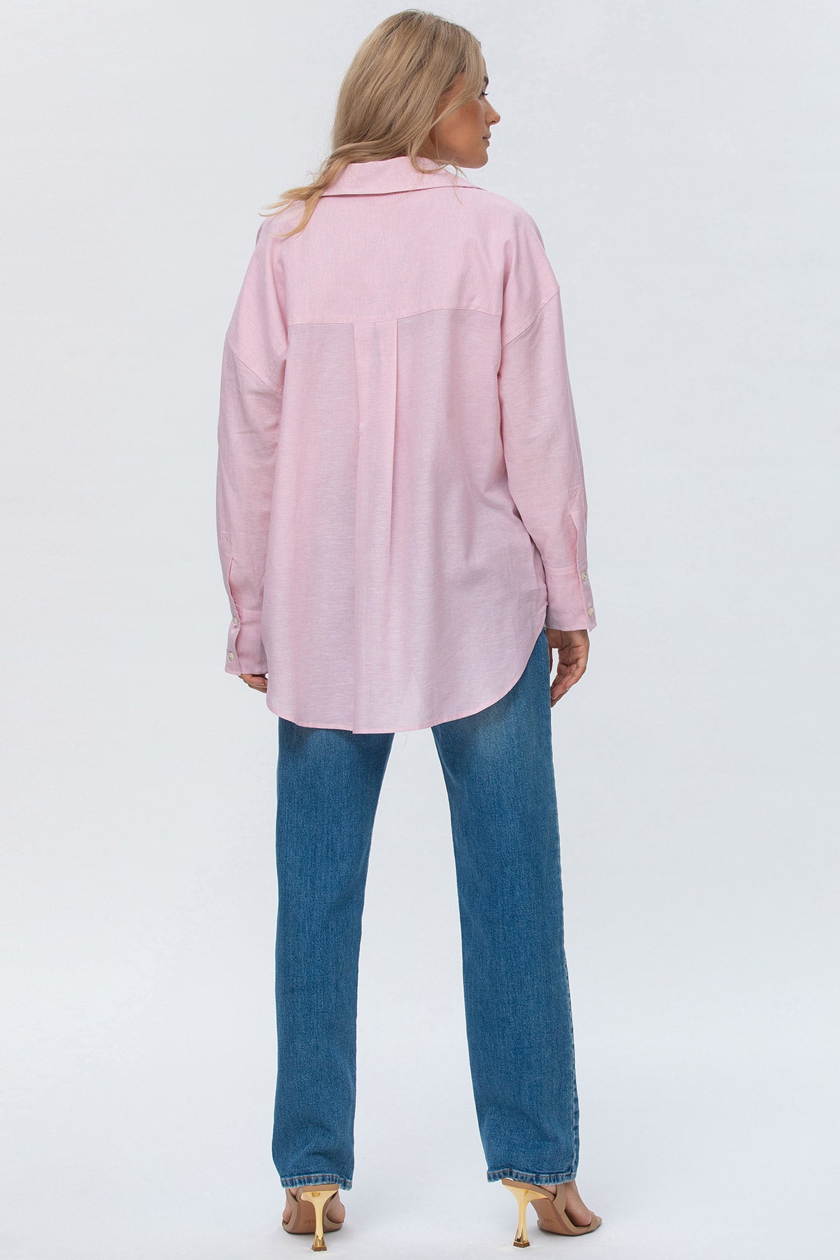 Donna di spalle con camicia rosa oversize e jeans dritti premaman, sfondo neutro.