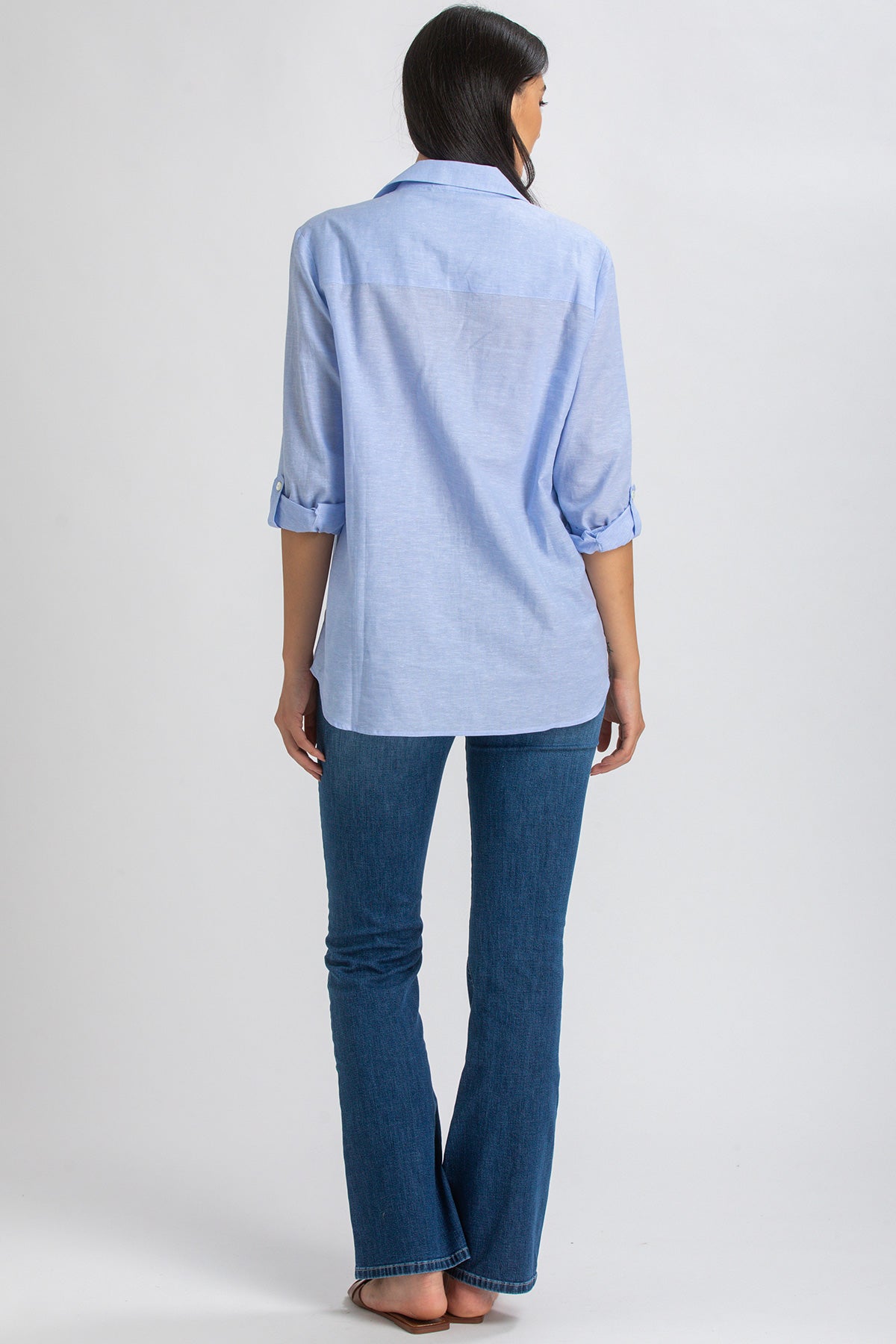 Donna di spalle indossa camicia premaman azzurra e jeans flare, sfondo neutro.