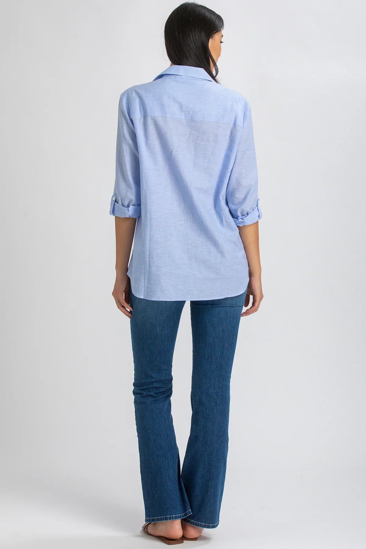 Donna di spalle indossa camicia premaman azzurra e jeans flare, sfondo neutro.