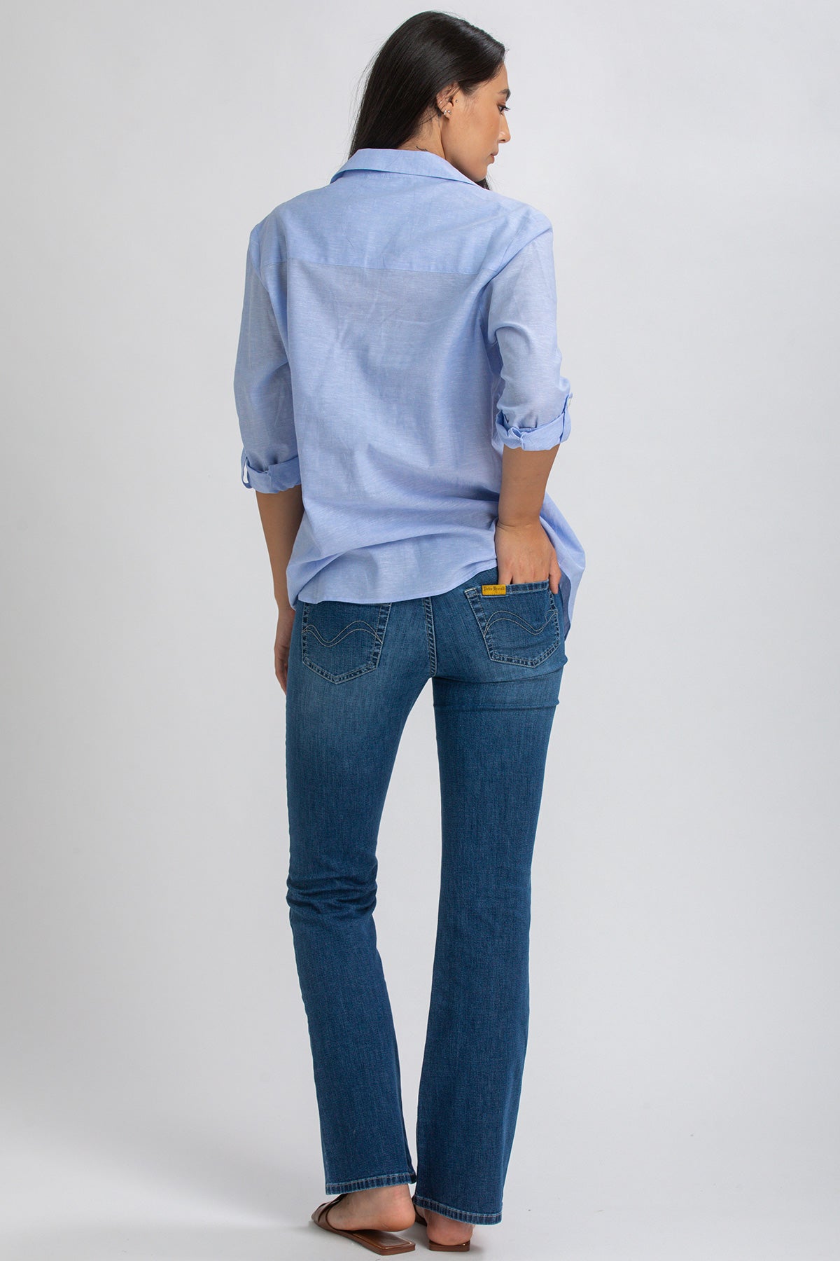 Donna di spalle con camicia azzurra e jeans premaman blu su sfondo neutro.