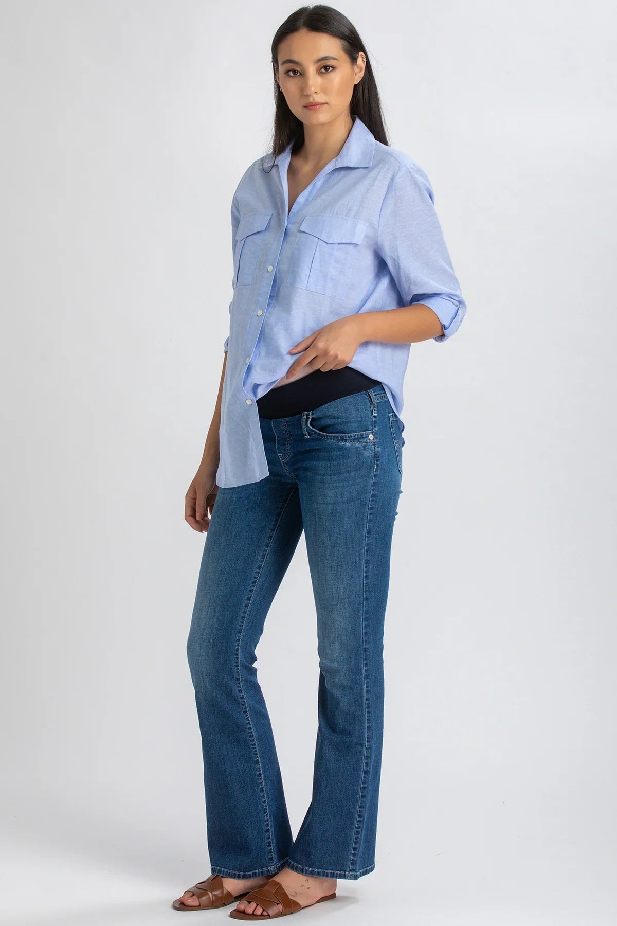 Donna incinta con camicia azzurra premaman e jeans flare in posa su sfondo neutro.