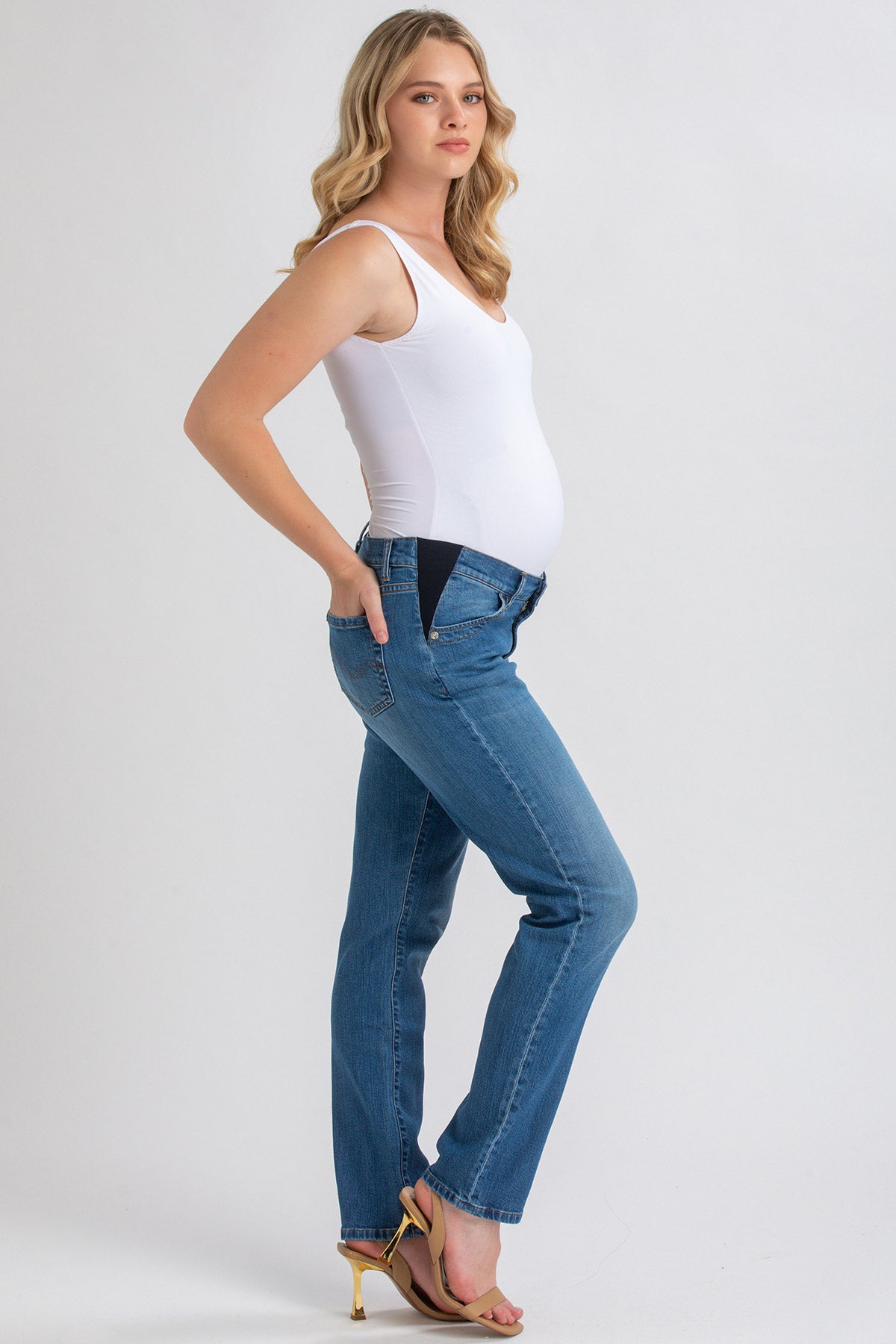 Donna incinta con jeans premaman a vita bassa e top bianco, posa su sfondo neutro