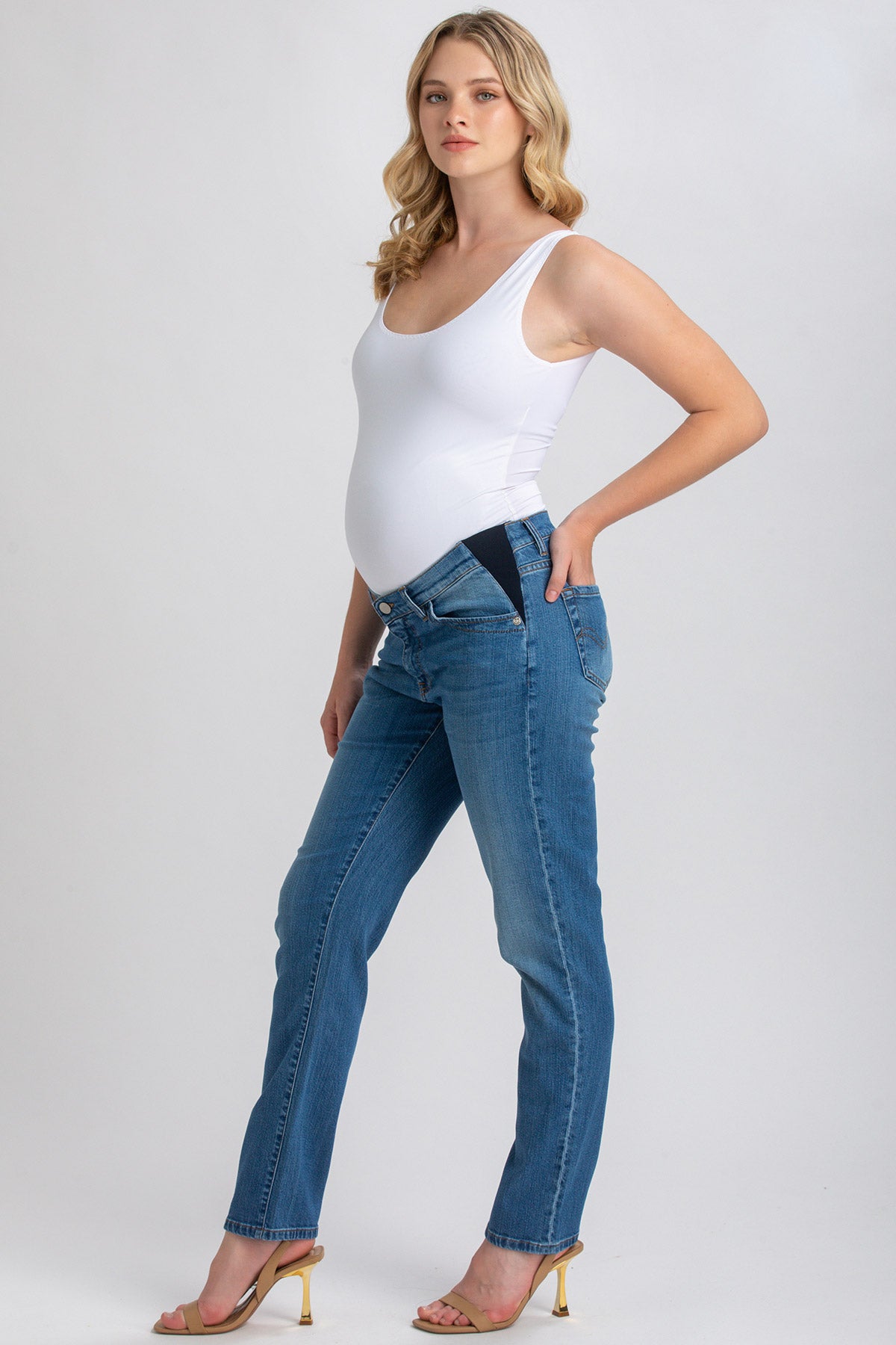 Modella incinta indossa jeans premaman elasticizzati e top bianco su sfondo neutro.