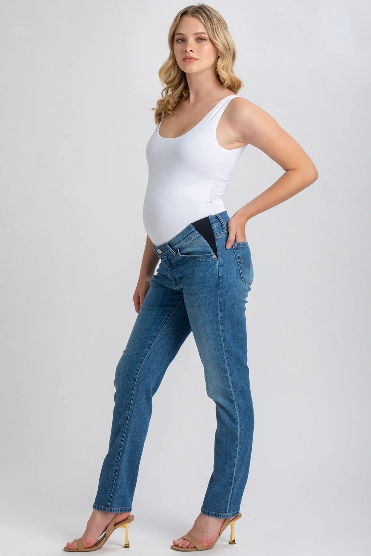 Modella incinta indossa jeans premaman elasticizzati e top bianco su sfondo neutro.