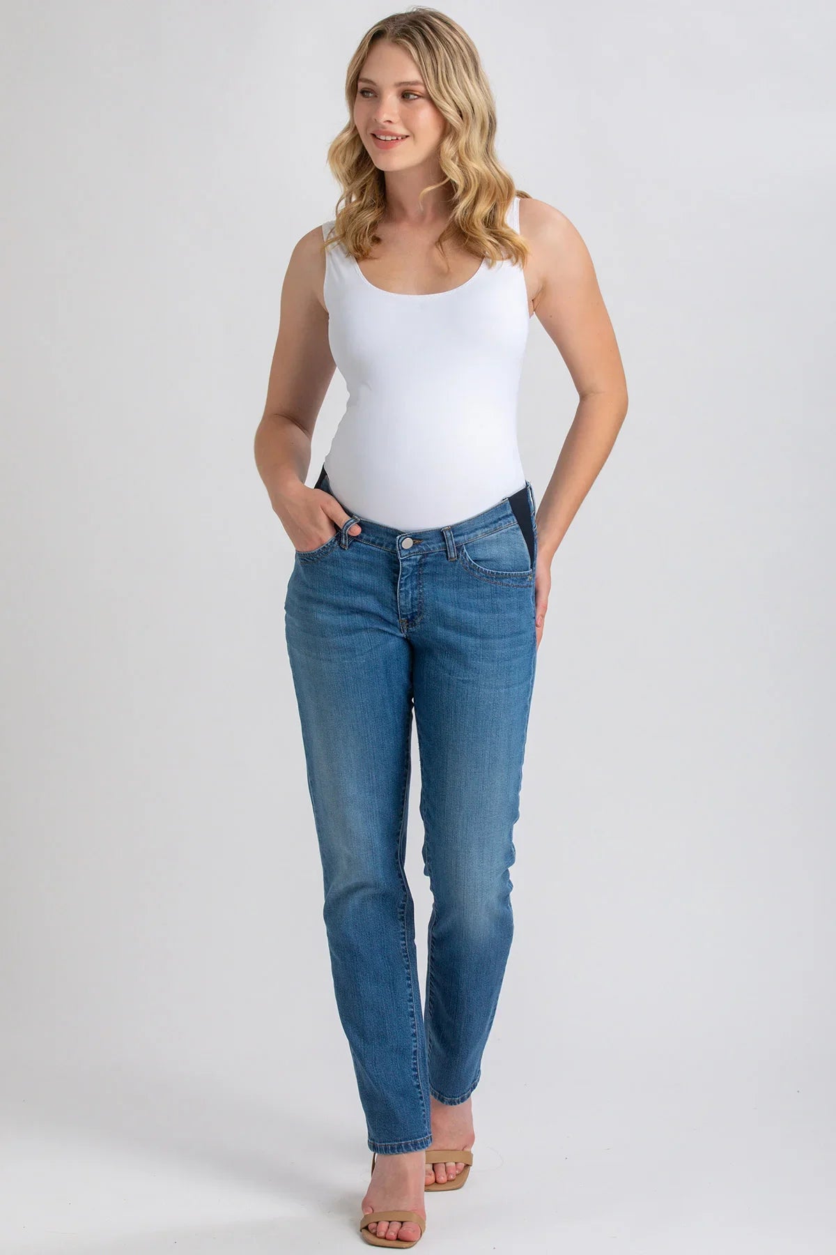 Donna incinta indossa jeans premaman elasticizzati blu e top bianco, su sfondo neutro.