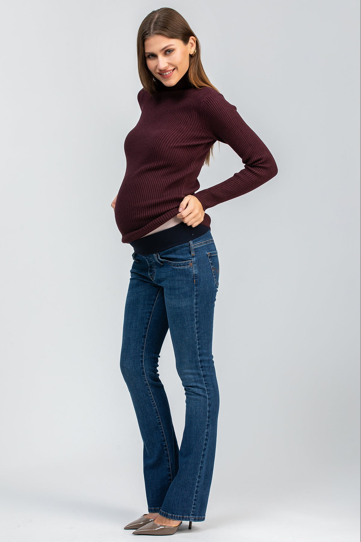 Modella incinta indossa jeans premaman bootcut blu e maglione bordeaux, fondo neutro.