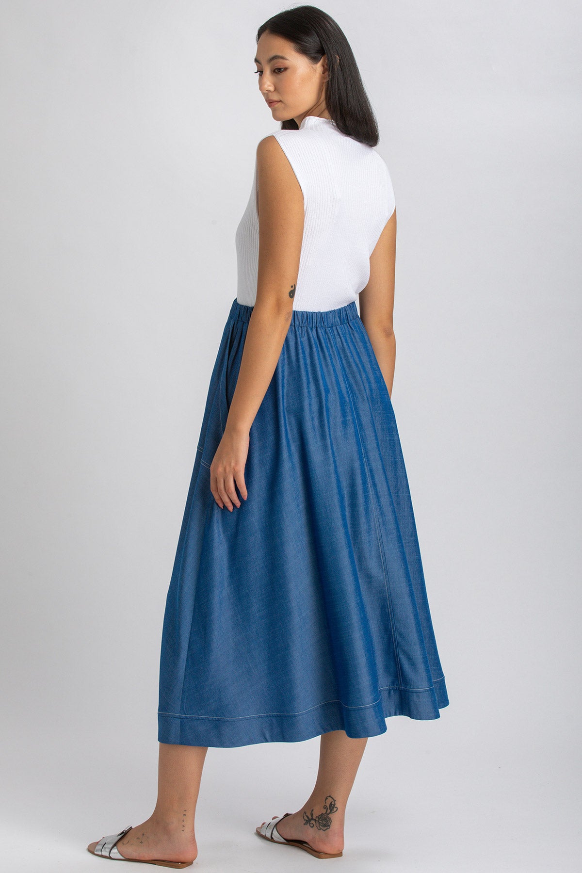 Donna con top bianco senza maniche e gonna lunga premaman in denim blu, vista di profilo.