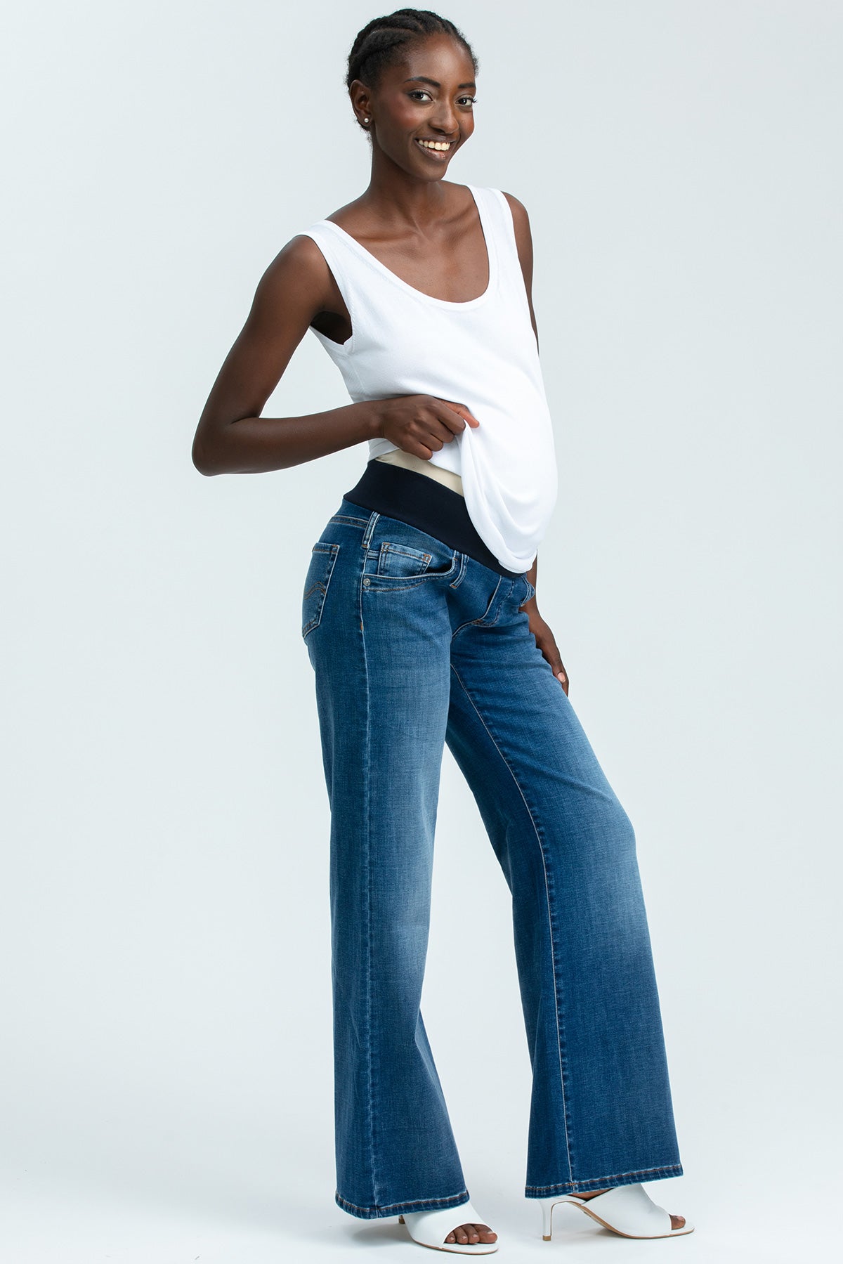 Donna incinta indossa jeans premaman a gamba larga in denim blu e top bianco, fondo bianco.