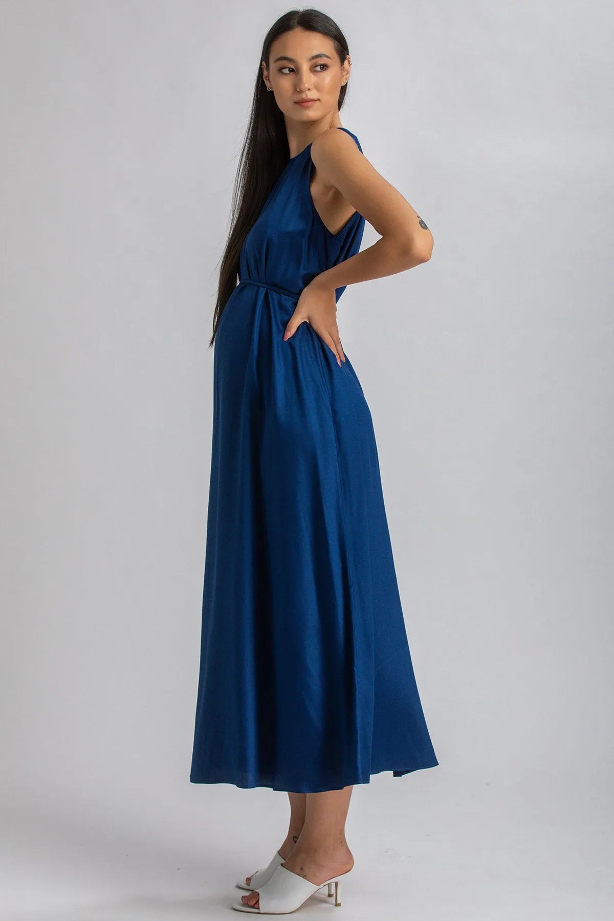Donna incinta indossa abito premaman lungo blu elegante con sandali bianchi, sfondo neutro.