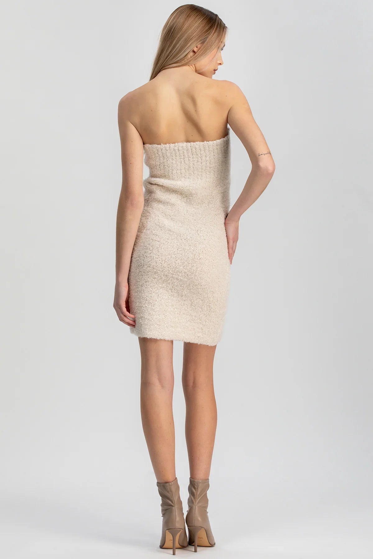 Donna di spalle con abito premaman corto senza spalline beige, stivaletti tacco alto, sfondo neutro.