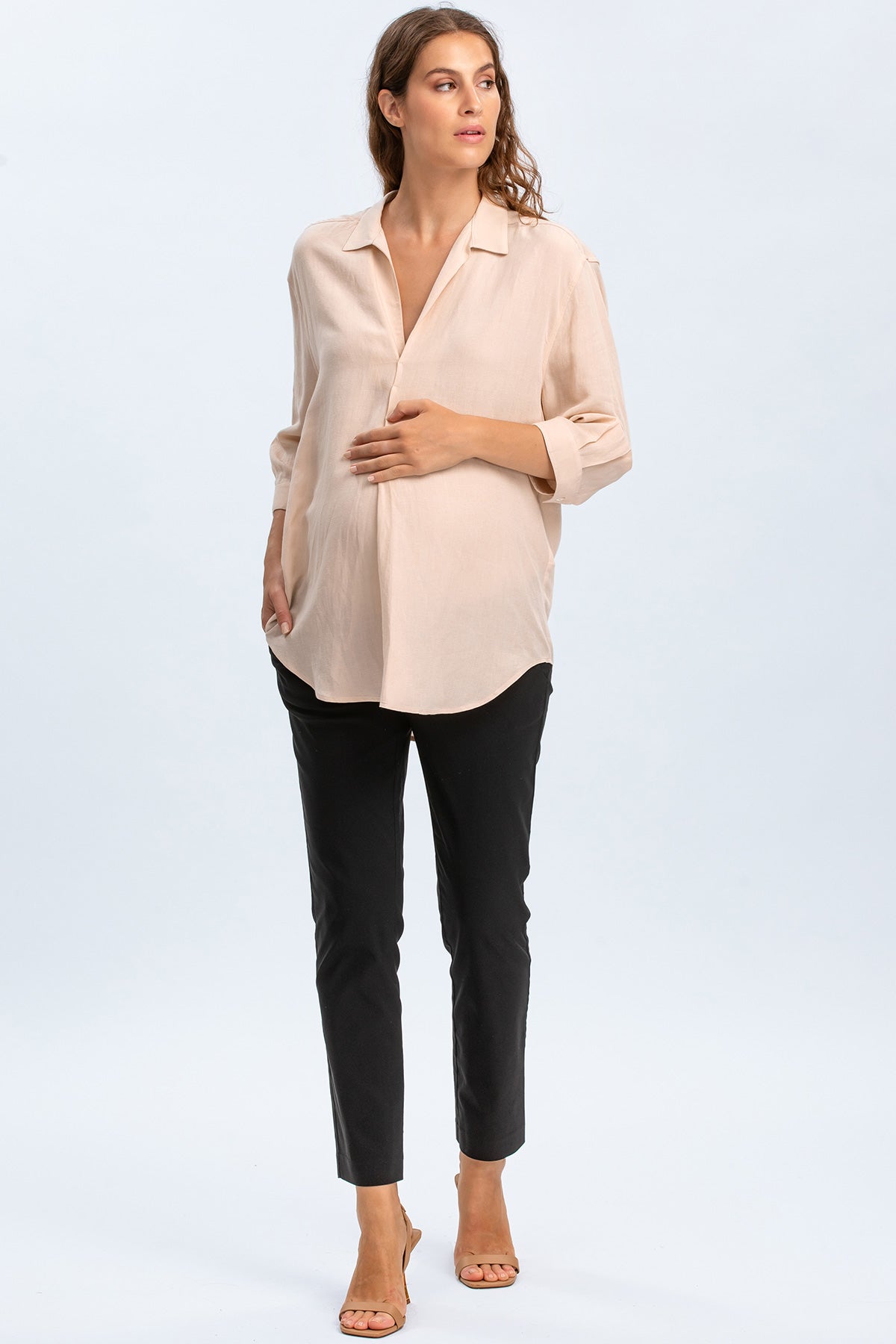 Donna incinta che indossa camicia premaman beige e pantaloni neri eleganti, fondo chiaro.