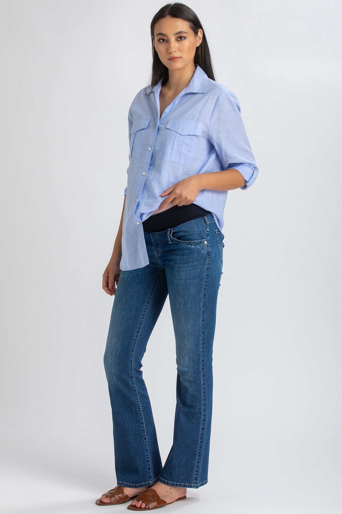 Donna incinta con camicia azzurra premaman e jeans flare in posa su sfondo neutro.