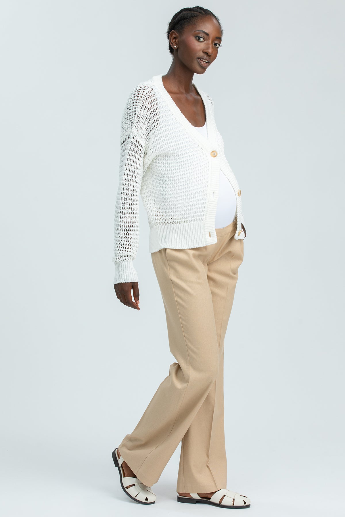 Modella incinta indossa cardigan premaman bianco traforato, pantaloni beige e sandali eleganti.