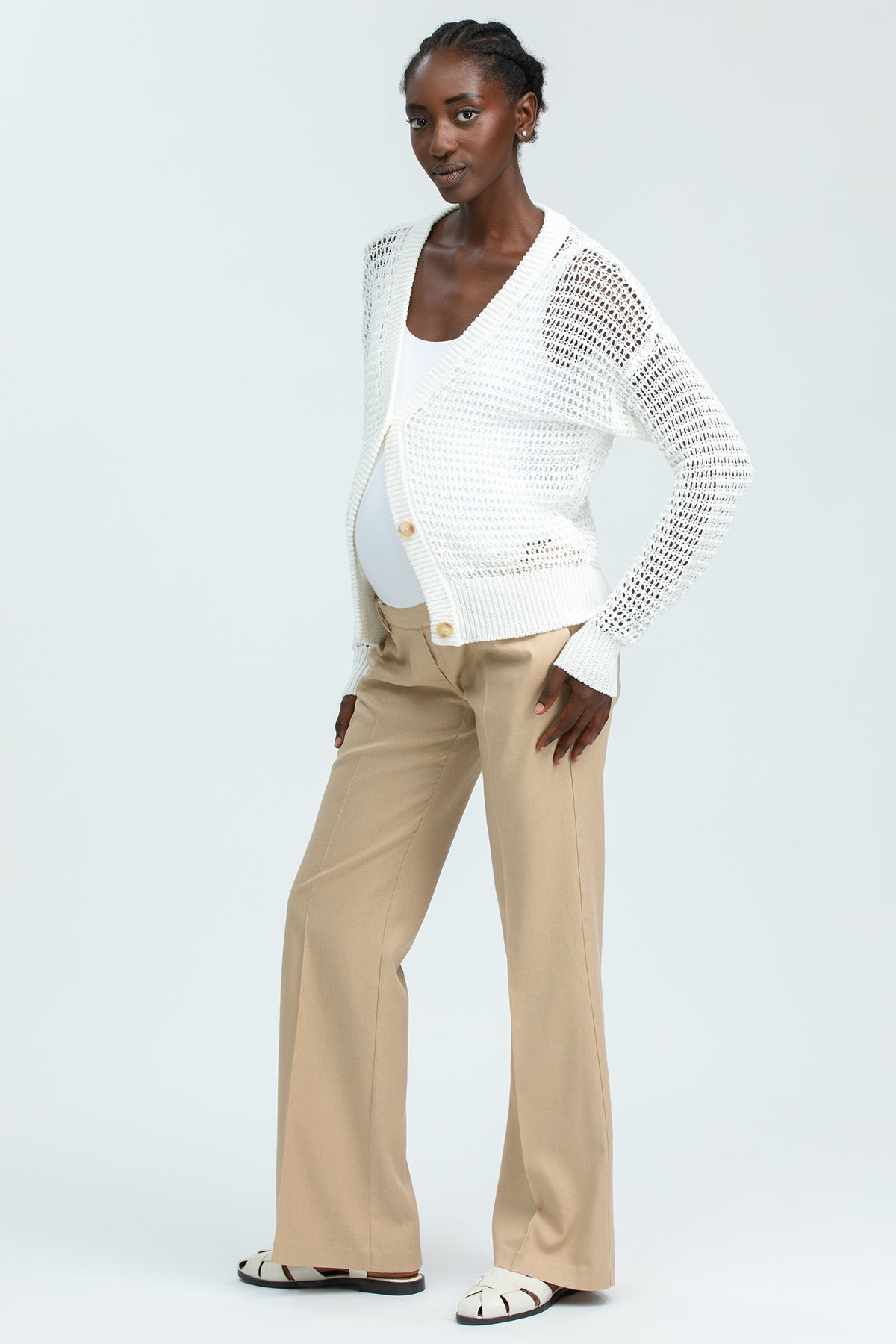 Modella premaman con pantaloni beige e cardigan traforato bianco su fondo neutro