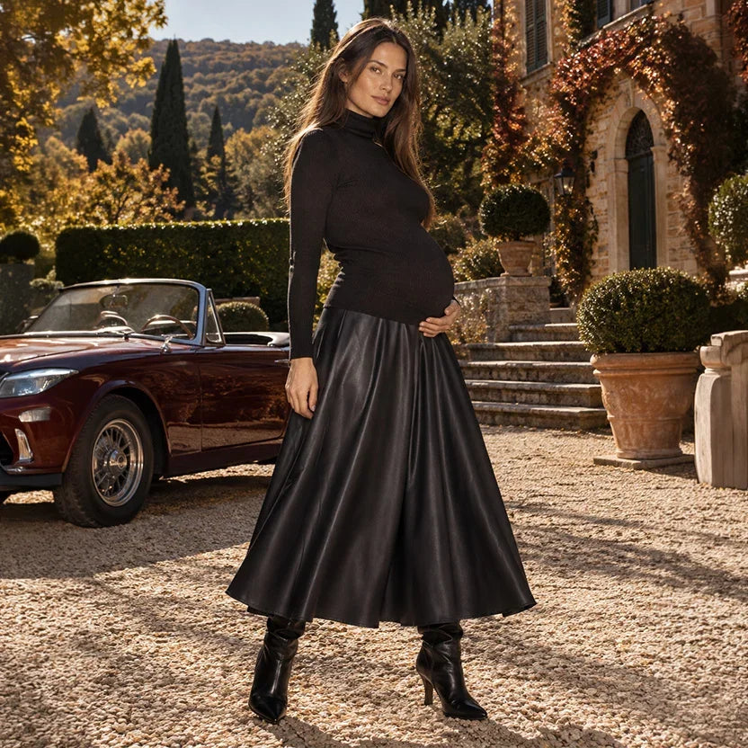 Donna incinta con maglia premaman nera a collo alto e gonna midi premaman effetto pelle, davanti a un'auto vintage nel cortile di una villa toscana.