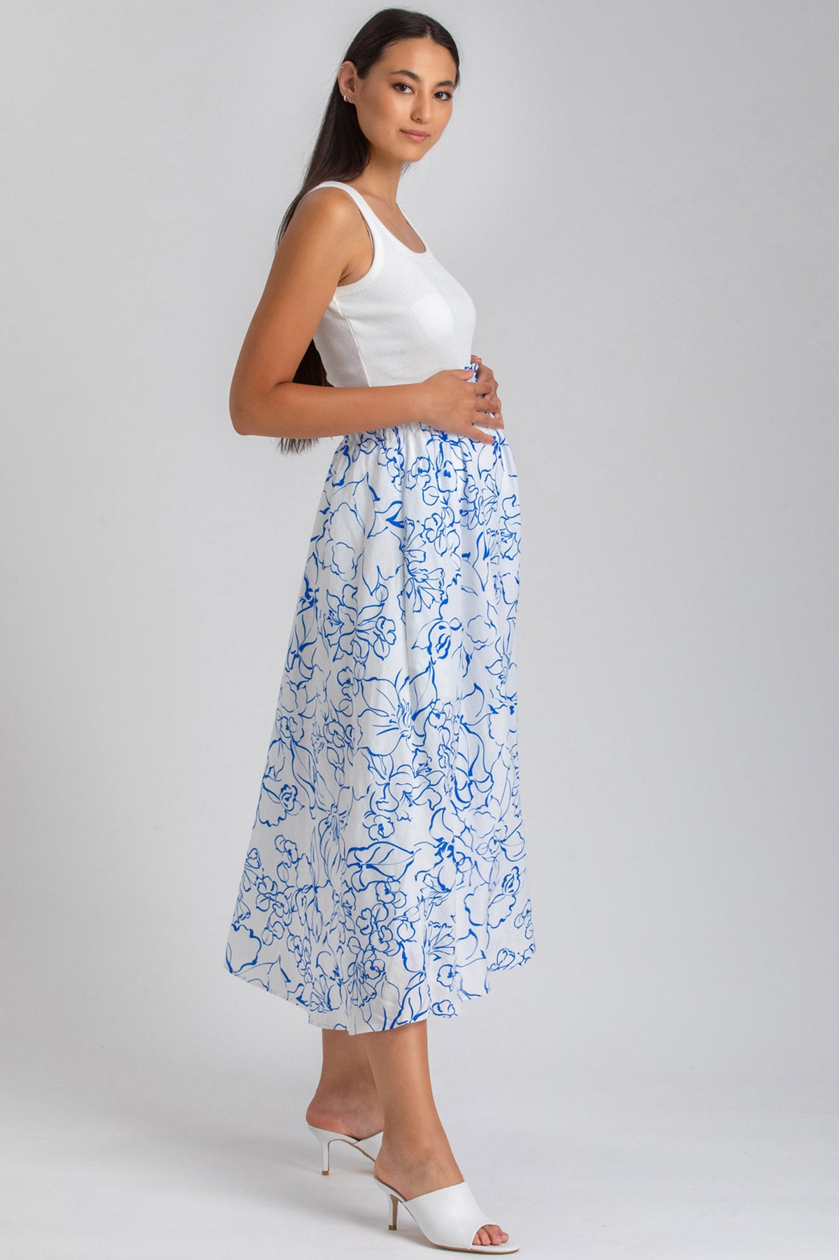 Donna incinta con abito premaman elegante bianco a fantasia floreale blu, su sfondo neutro.