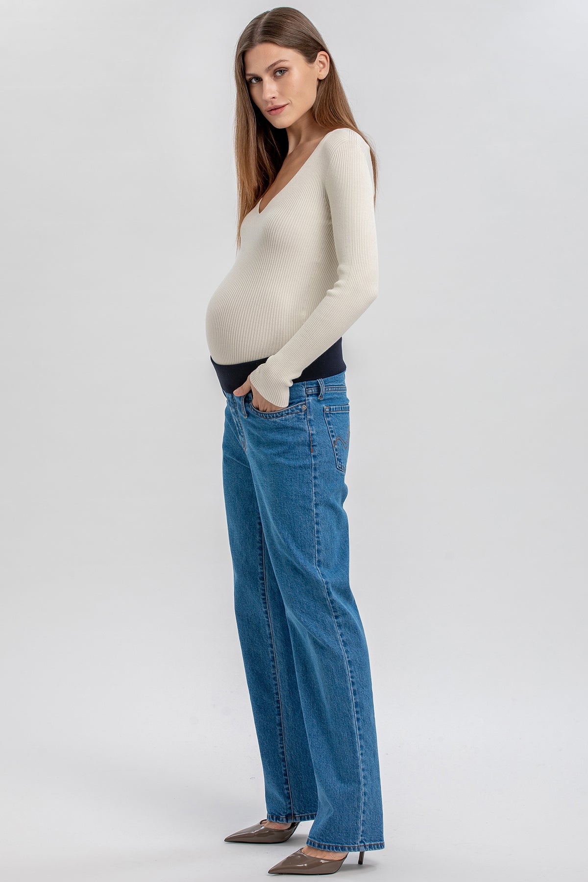 Modella incinta con maglia beige e jeans premaman a gamba dritta su sfondo neutro.