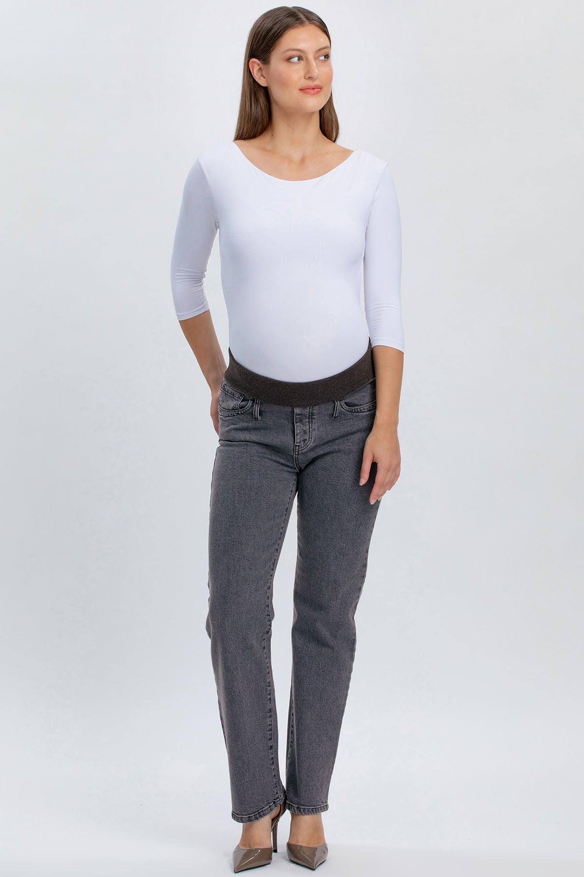 Donna incinta con jeans premaman grigi a gamba dritta e maglia bianca, su sfondo neutro.