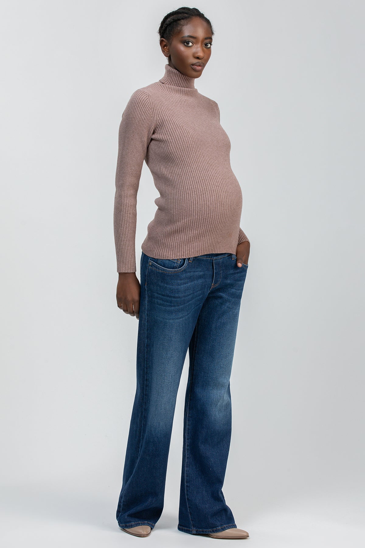Modella incinta con maglione premaman a collo alto beige e jeans flare, fondo neutro.