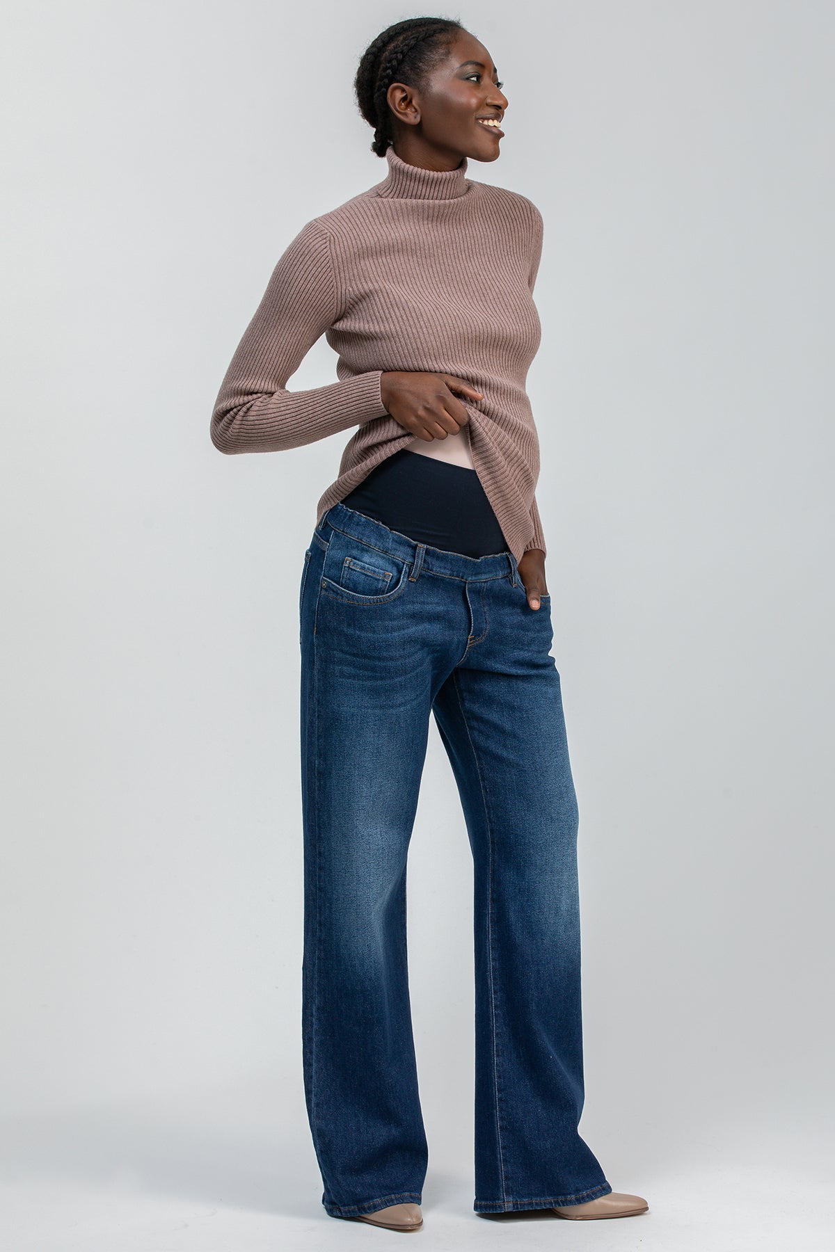 Modella indossa jeans premaman blu a gamba larga e maglione a collo alto rosa, su sfondo neutro.