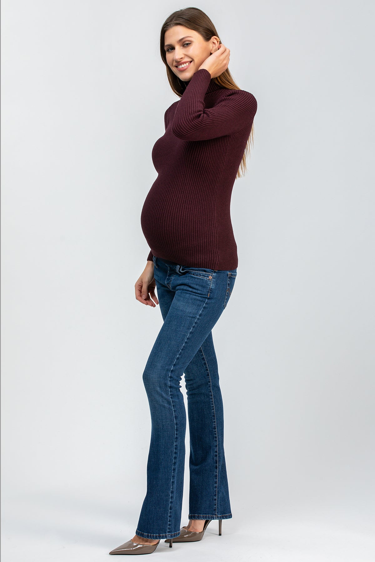 Donna incinta indossa jeans premaman slim bootcut e maglione bordeaux, sfondo neutro.
