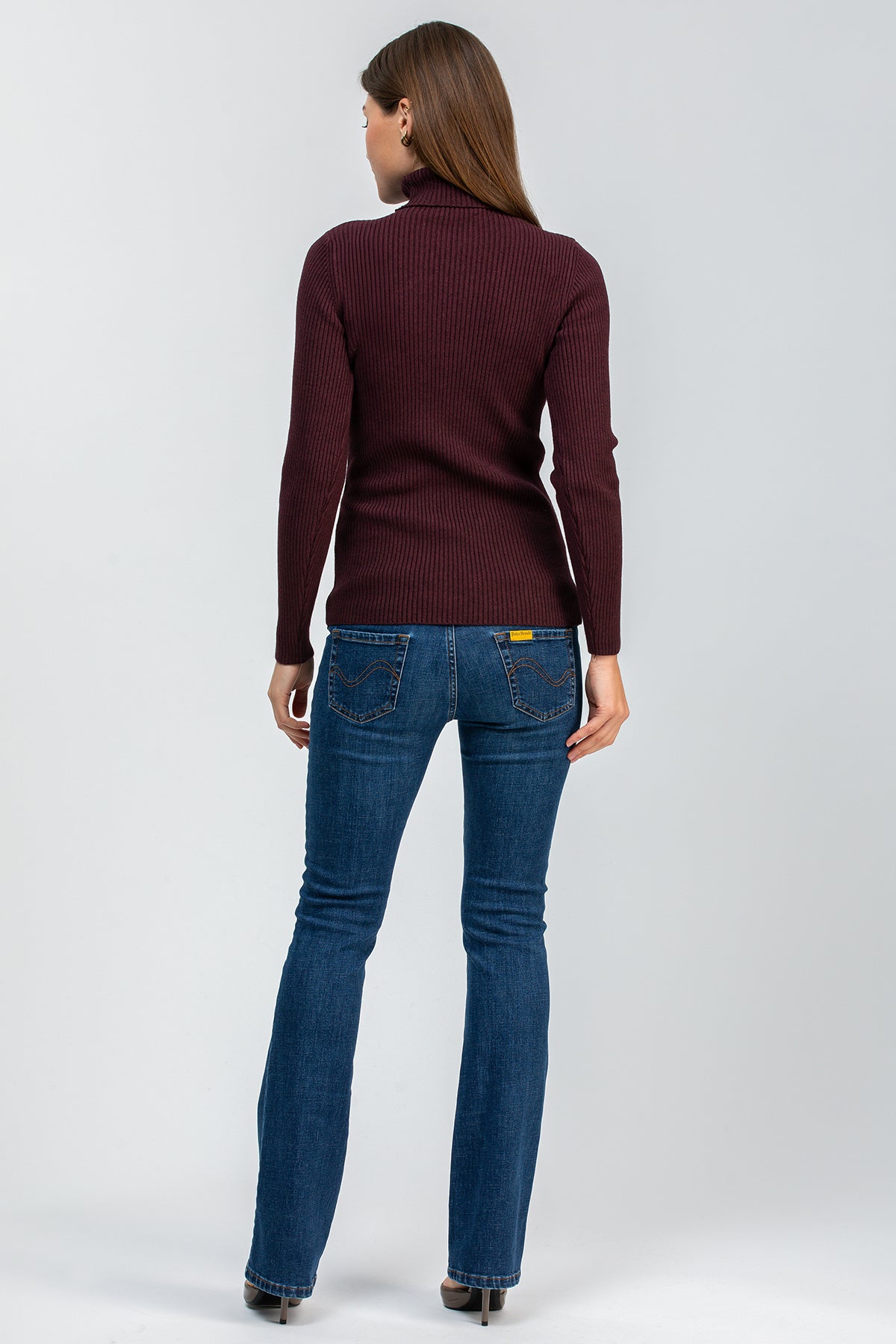 Donna di spalle con maglione bordeaux e jeans premaman bootcut blu, su sfondo neutro