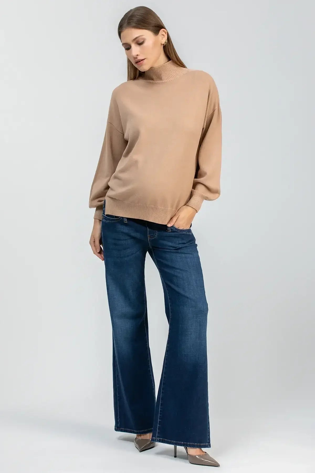 Modella indossa maglione premaman beige a collo alto e jeans a zampa, look casual elegante.