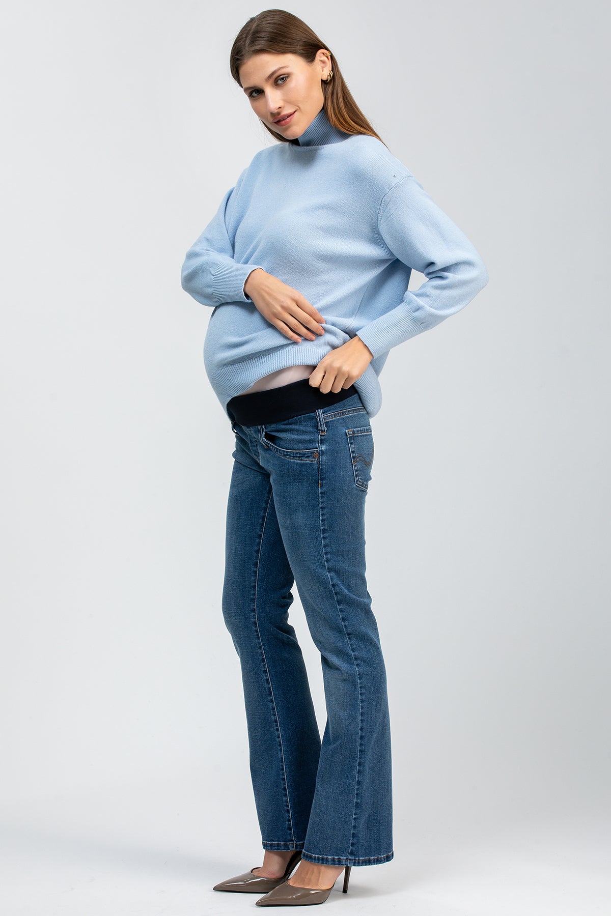 Donna incinta con maglione azzurro e jeans premaman flare, su sfondo neutro.