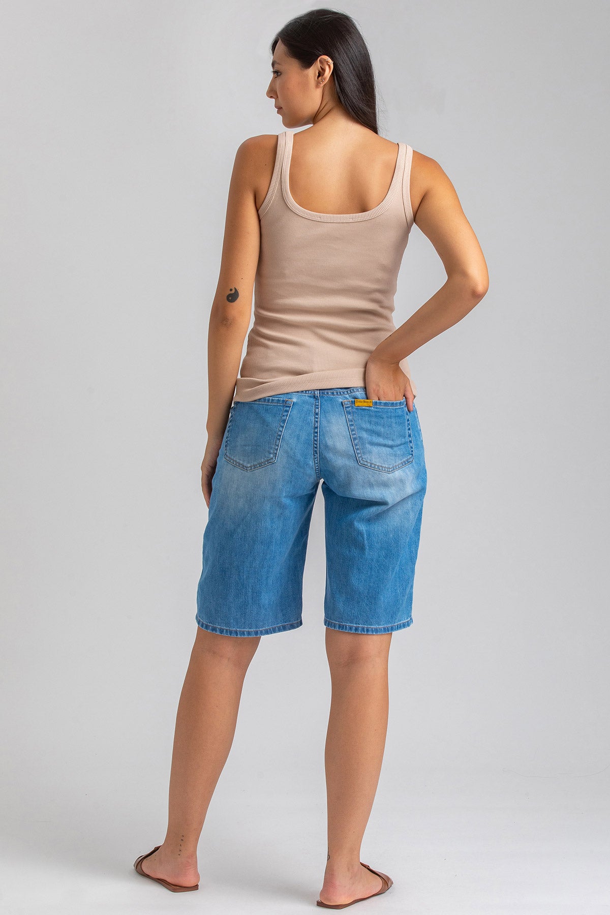 Donna di spalle indossa bermuda premaman in jeans e canotta beige, sfondo neutro