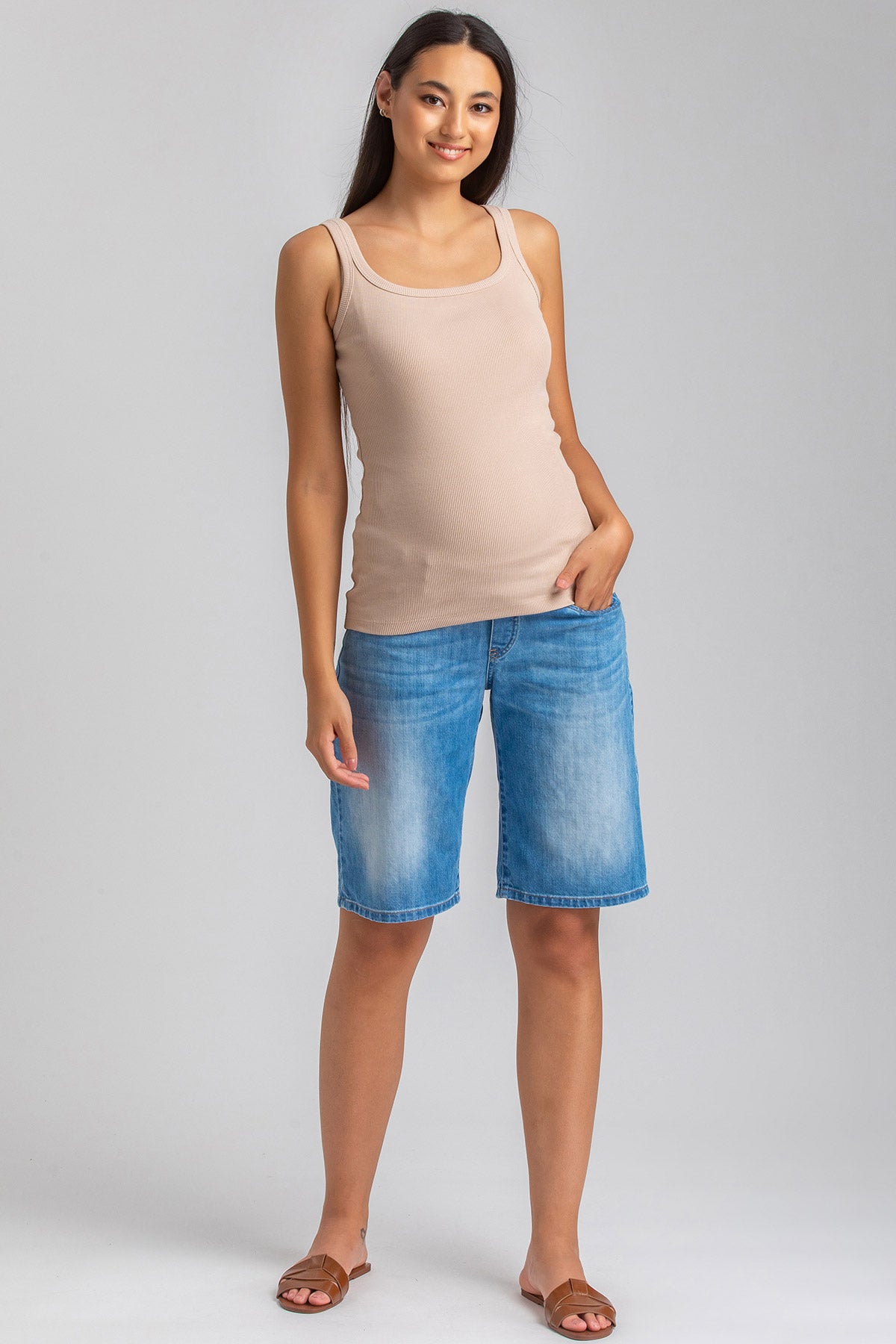 Donna incinta con canotta beige e bermuda premaman in denim chiaro, sfondo neutro.