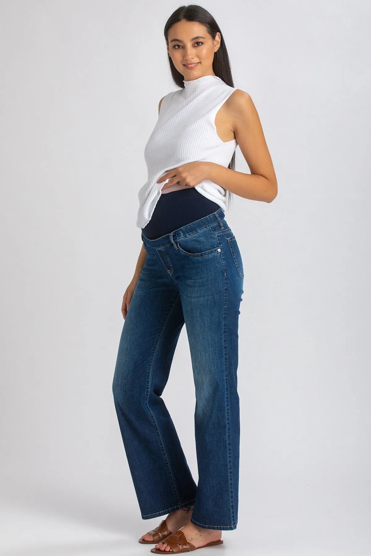 Donna incinta con jeans premaman a vita alta e top bianco senza maniche su sfondo neutro.