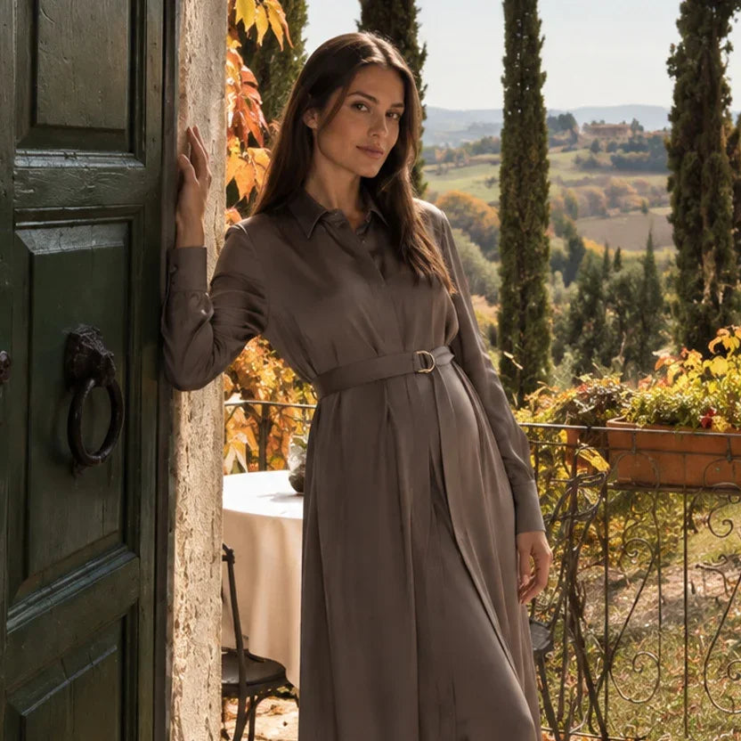 Donna incinta con abito premaman lungo stile chemisier color tortora, con maniche lunghe e cintura in vita, davanti a una villa toscana.