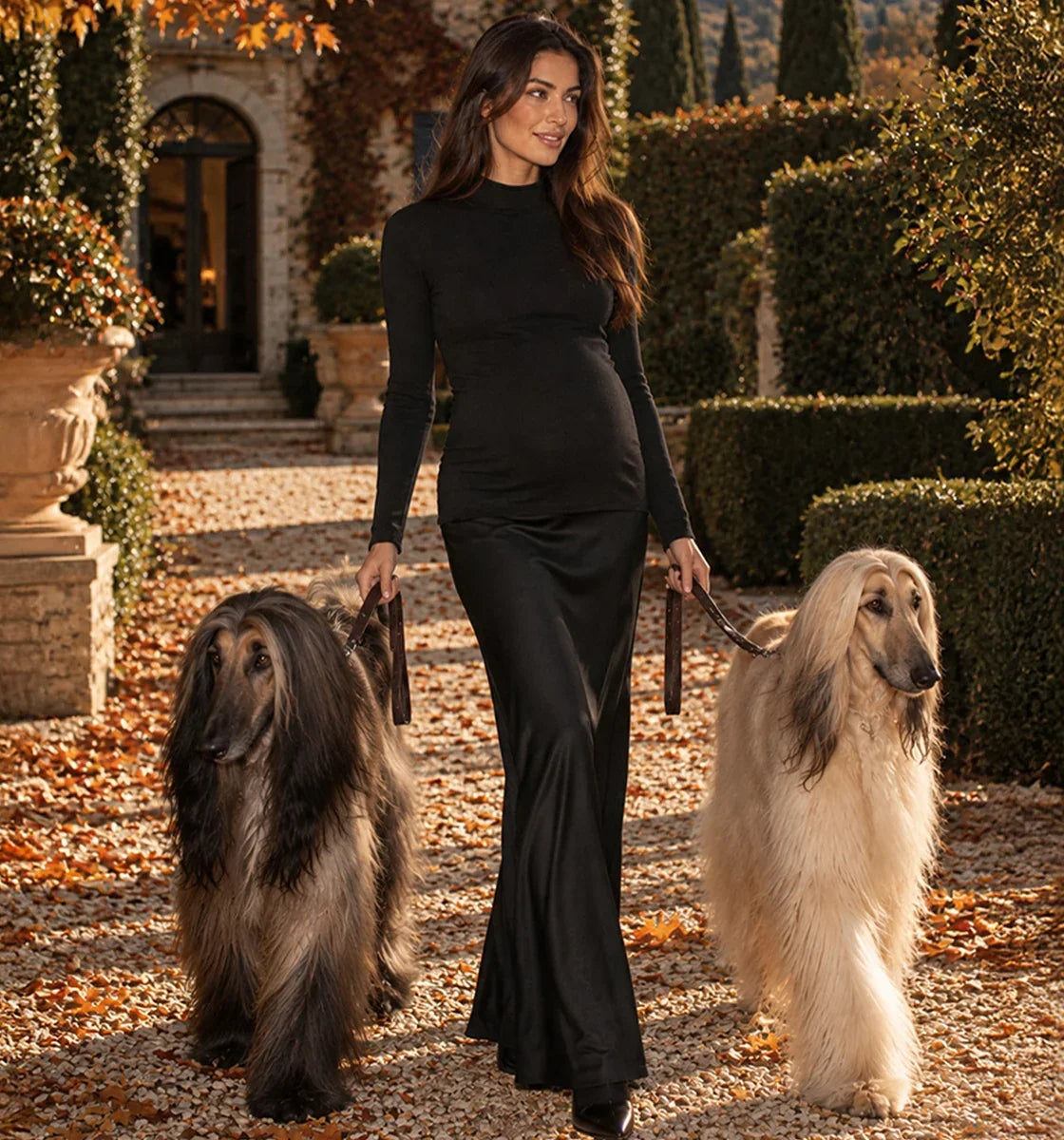 Donna con abito premaman nero elegante che passeggia con due levrieri afghani in un giardino.